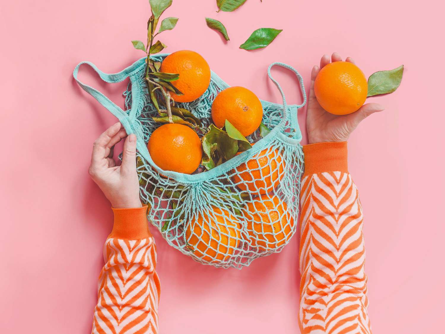 Woman hands in orange striped sweatshirt holding oranges with green leaves in blue eco-friendly shopping mesh bag on pink background.